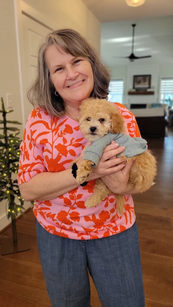 A smiling woman holds a small, fluffy puppy dressed in a blue sweater, standing in a well-lit living room with a decorative tree in the background.